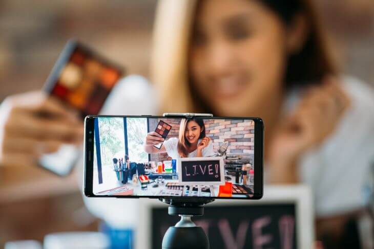 Happy young attractive Asian ethnic model filming vlog on mobile phone on tripod and showing eyeshadows at table with cosmetics