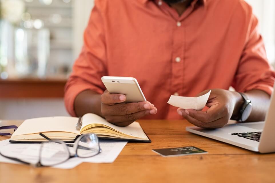 Closeup of man's hands using phone to calculate expenses. Man checking invoice balance on mobile phone app. Close up hands of black guy taking a closer look at his budget and calculating credit card bills.